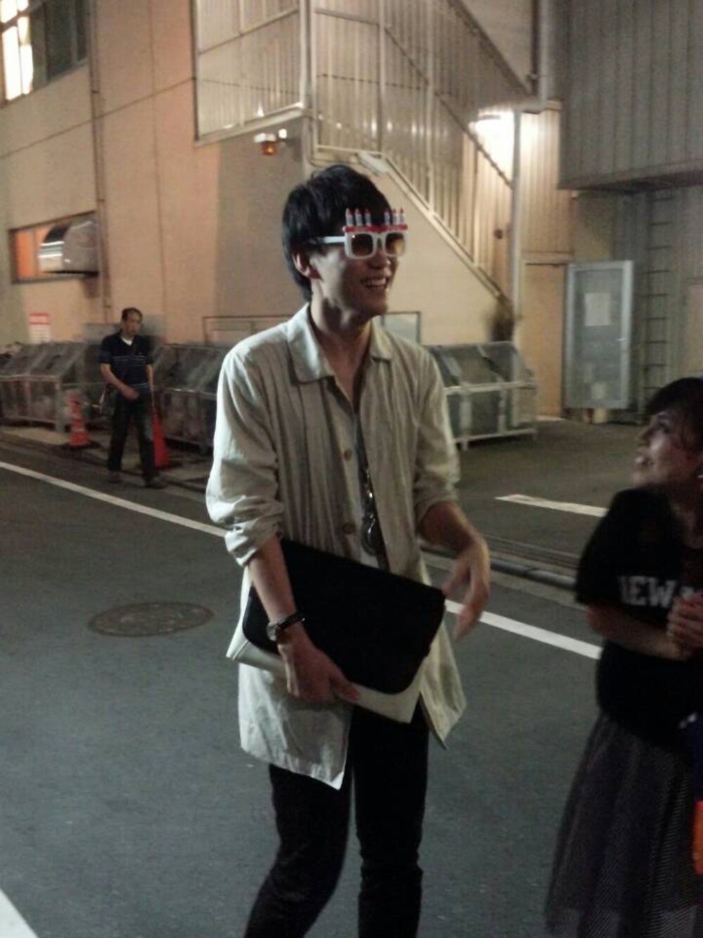 ベージュ系のステンカラーコート、シルバー系のネックレス、ブラック系のクラッチバッグを着用したメンズの夏コーディネートの1枚目の写真