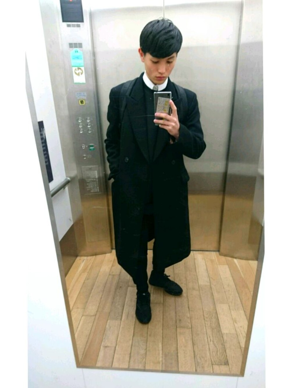 ブラック系のチェスターコート、ブラック系のスニーカー、ブラック系のTシャツ/カットソーを着用したメンズの冬コーディネートの1枚目の写真