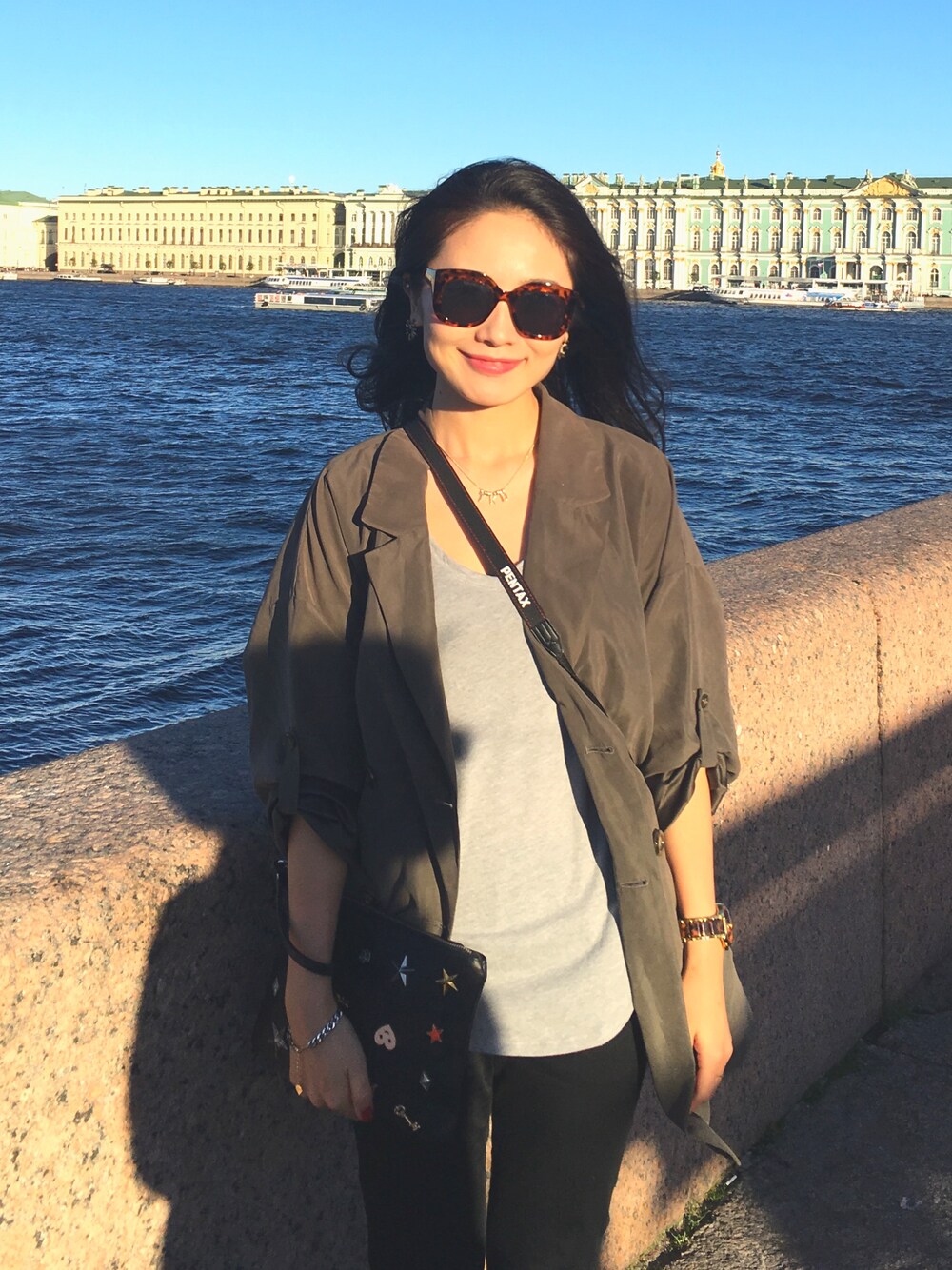 グリーン系のトレンチコート、グレー系のTシャツ/カットソー、ブラック系のクラッチバッグを着用したレディースの秋コーディネートの1枚目の写真