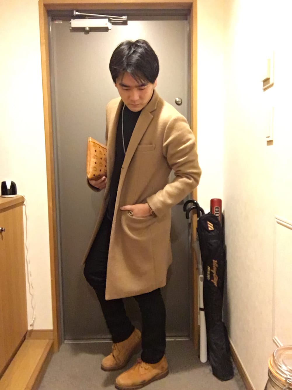 ベージュ系のチェスターコート、ブラック系のスウェット、ブラウン系のクラッチバッグを着用したメンズの冬コーディネートの1枚目の写真